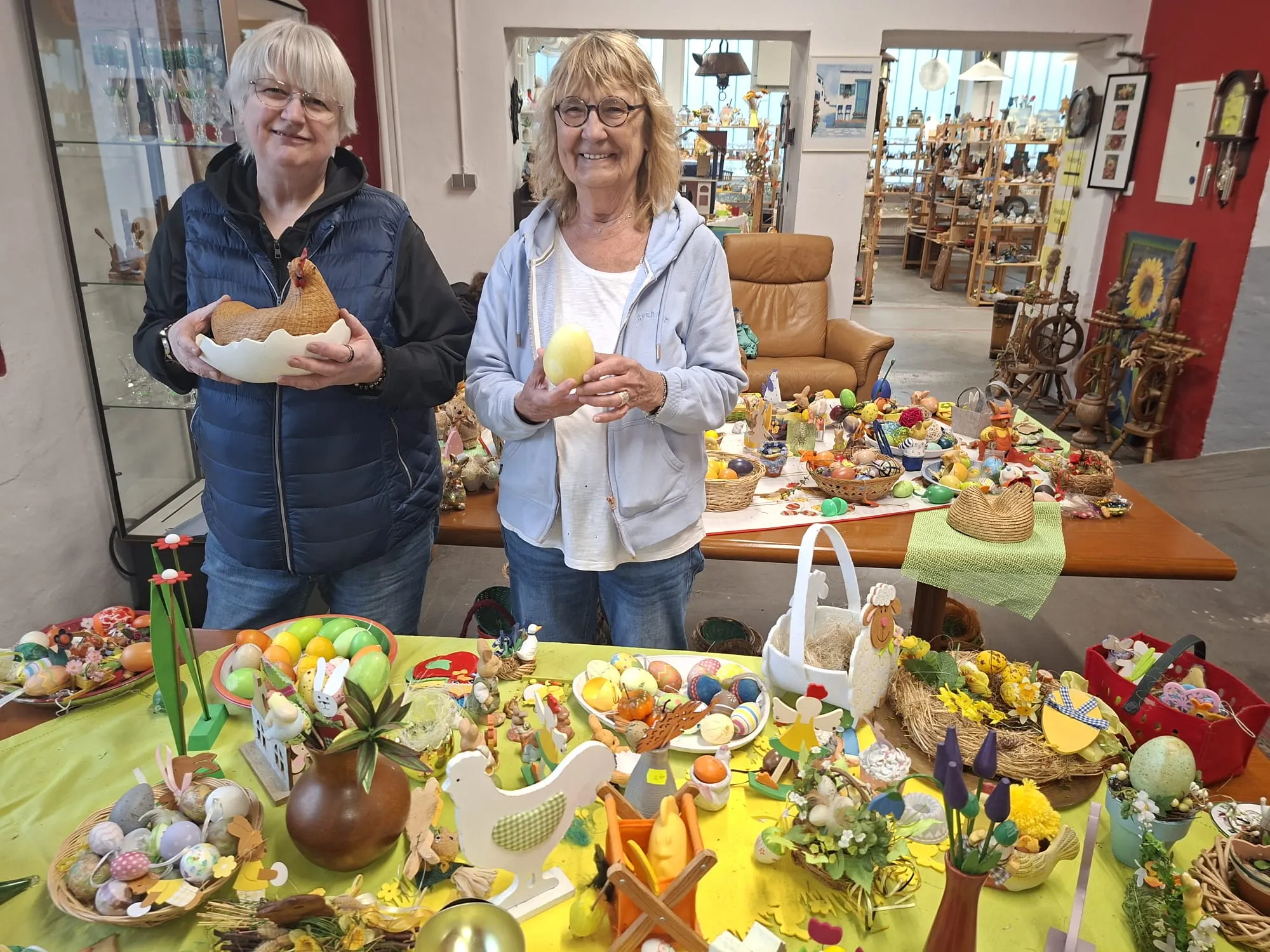 Zwei Frauen stehen vor einem reich gedeckten Tisch mit zahlreichen bunten Ostereiern und Dekorationen. Im Hintergrund sind weitere Möbel und Ausstellungsstücke in einem Geschäft zu sehen.