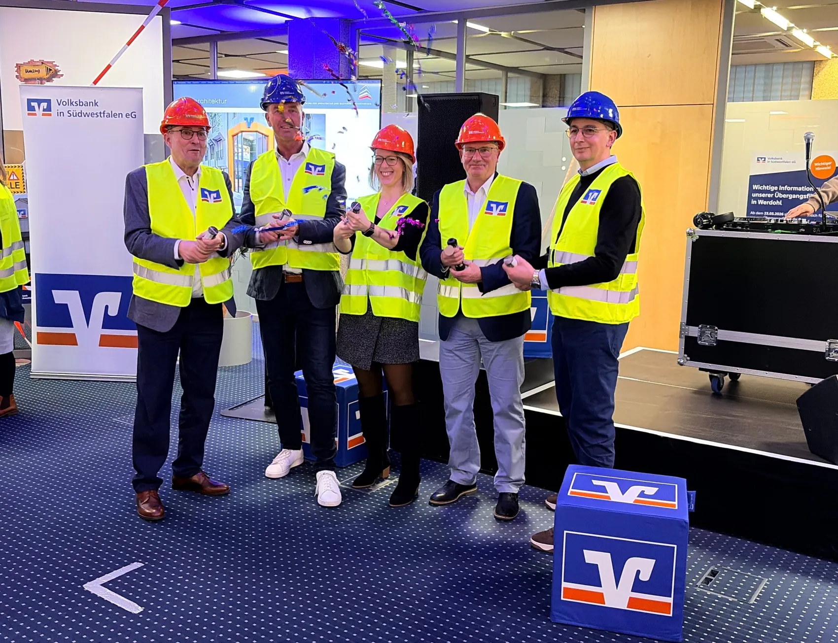 Fünf Personen in Schutzhelmen und Warnwesten stehen vor einer Werbetafel der Volksbank und halten symbolisch Bänder. Im Hintergrund ist die Baustelle der Filiale in Werdohl zu sehen.
