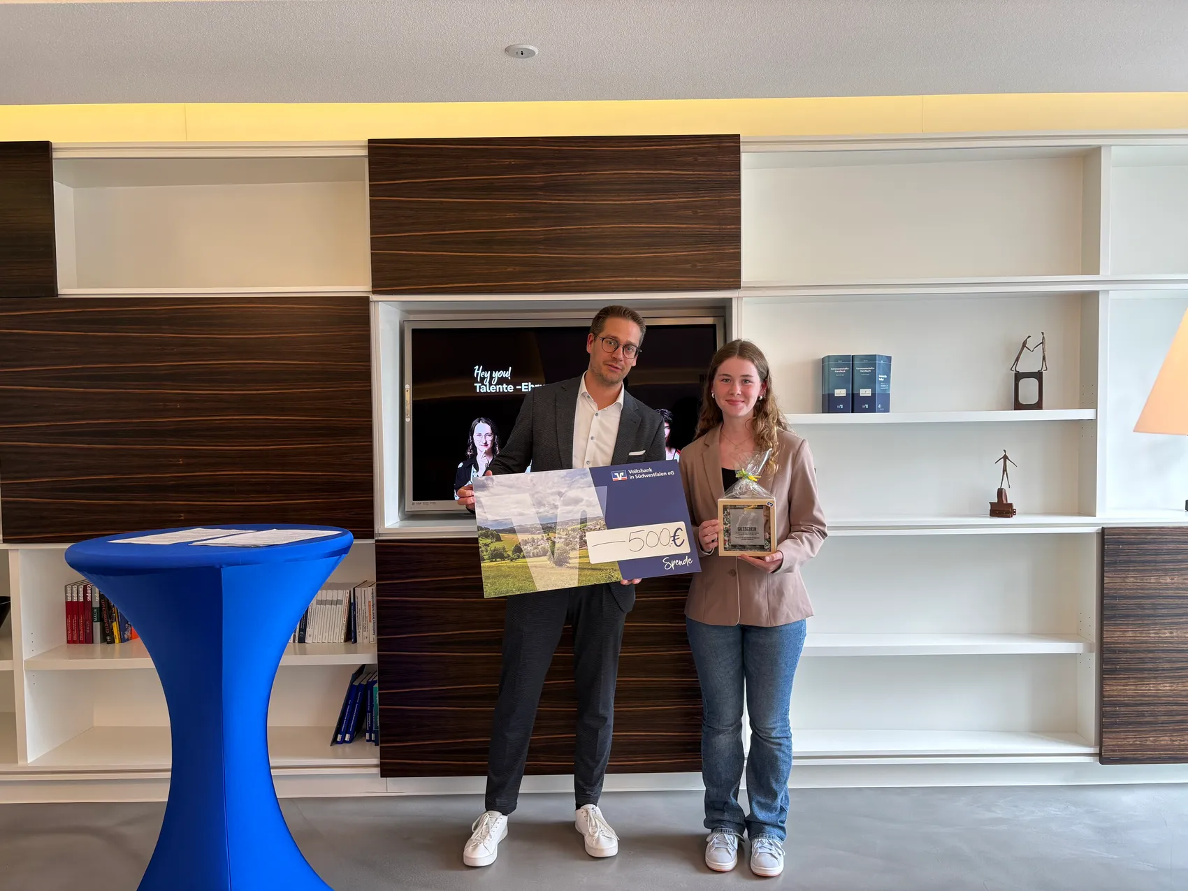 Ein Mann und ein Mädchen stehen nebeneinander und halten einen Scheck über 500 Euro und eine Auszeichnung. Im Hintergrund ist ein Bildschirm mit einer Person darauf zu sehen.