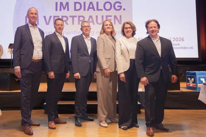 Fünf Personen stehen auf einer Bühne vor einem Hintergrund mit der Aufschrift „IM DIALOG. VERTRAUEN“. Die beiden mittleren Personen, eine Frau in einem hellen Hosenanzug und ein Mann in einem dunklen Anzug, werden im Rahmen der Vertreterversammlung der Volksbank in Südwestfalen als designierte Vorstandsmitglieder vorgestellt.