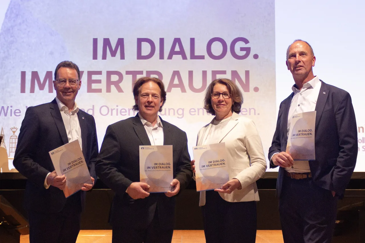 Vier Personen – zwei Männer und zwei Frauen – stehen auf einer Bühne und halten jeweils ein Buch mit dem Titel „IM DIALOG. IM VERTRAUEN.“ in den Händen. Im Hintergrund ist ein Banner mit demselben Schriftzug zu sehen.