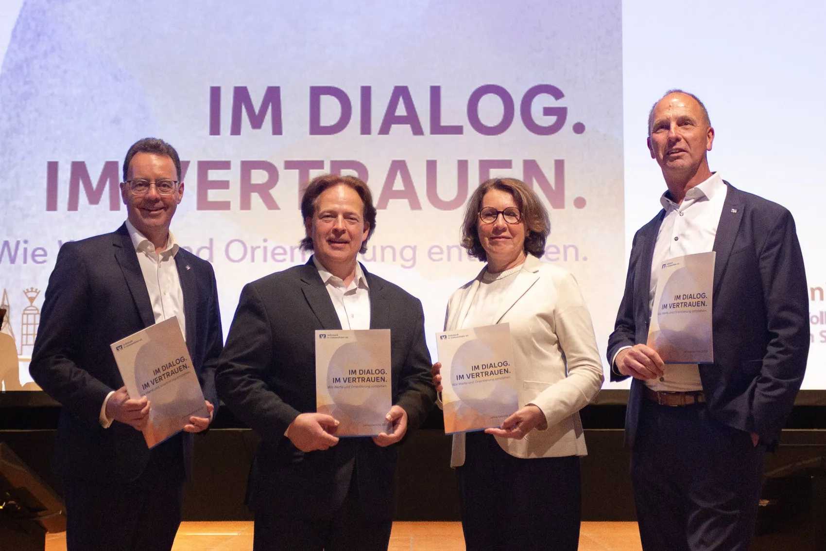 Vorstandssprecher Roland Krebs, Aufsichtsratsvorsitzender Prof. Dr.-Ing. Sven Keller, stellvertretende Aufsichtsratsvorsitzende Brigitta Wallmann und Vorstand Jens Brinkmann halten Informationsmaterialien während der Vertreterversammlung der Volksbank in Südwestfalen. Im Hintergrund ist das Motto „IM DIALOG. IM VERTRAUEN.“ zu sehen.