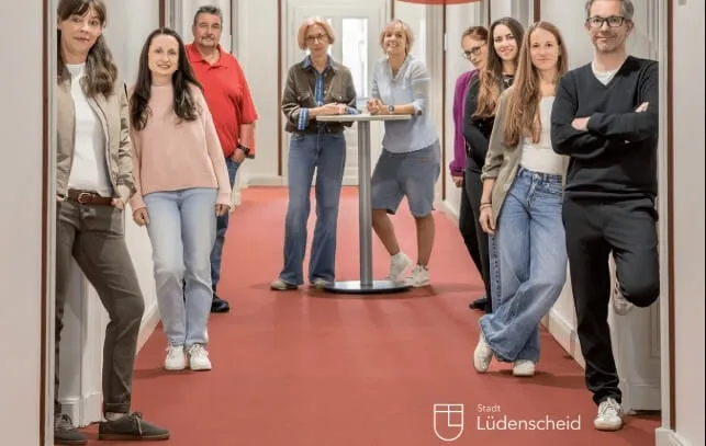 Eine Gruppe von Menschen steht lächelnd in einem hellen Flur, darunter Michael Tschöke (rechts). Im Hintergrund ist das Logo der Stadt Lüdenscheid zu sehen.