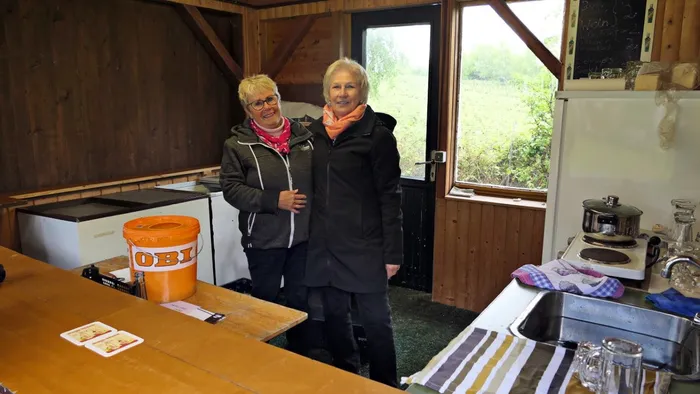 Zwei Frauen stehen in einem hellen Raum mit Holzwandverkleidung, vermutlich in einem Wanderheim. Im Hintergrund ist ein Fenster zu sehen.
