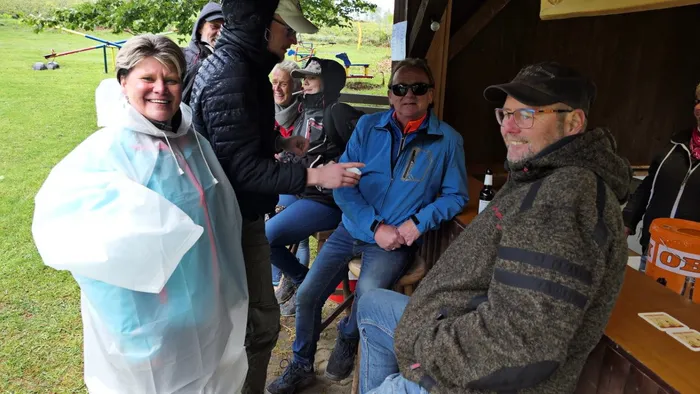 Mehrere Wanderer sitzen und stehen vor einem Unterstand, einige unterhalten sich und lächeln. Im Hintergrund eine Wiese und weitere Personen.