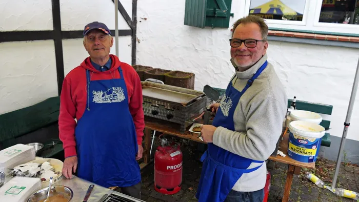Zwei Männer mit roten Pullovern und blauen Schürzen stehen lächelnd an einem Grill im Freien. Im Hintergrund ein Fachwerkhaus und ein Tisch mit Lebensmitteln.