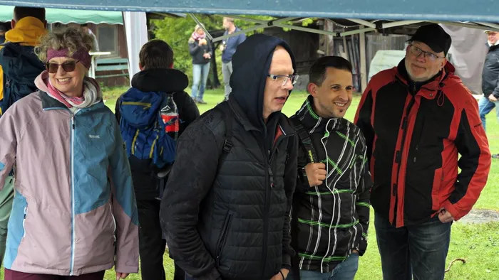 Vier Wanderer stehen unter einem Zelt und unterhalten sich. Im Hintergrund sind weitere Personen und ein grüner Rasen erkennbar.
