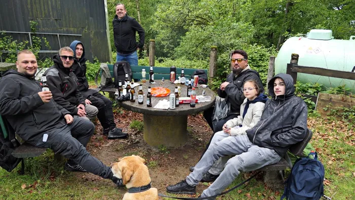 Eine Gruppe von Männern und Jugendlichen sitzt um einen Tisch im Freien, auf dem Getränke stehen. Im Hintergrund steht ein Mann und ein großer Gastank.