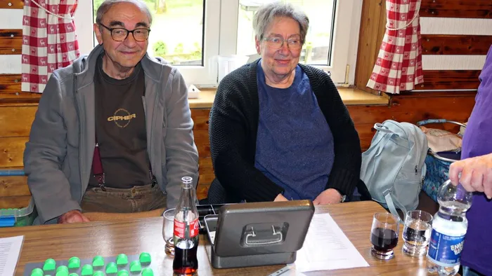Zwei ältere Menschen sitzen an einem Tisch mit Getränken und einem Laptop, im Hintergrund eine Wand mit rot-weißem Karomuster.