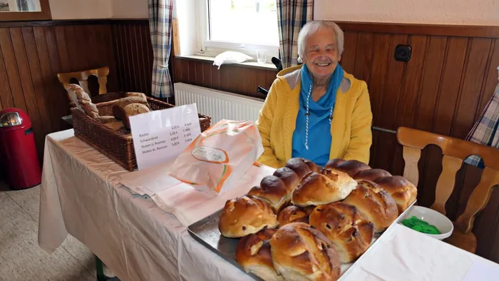Ältere Frau mit hellen Haaren lächelt hinter einem Tisch mit selbstgebackenen Waren, darunter Brot und Brötchen. Im Hintergrund ist ein rustikaler Holzwandpaneel zu sehen.