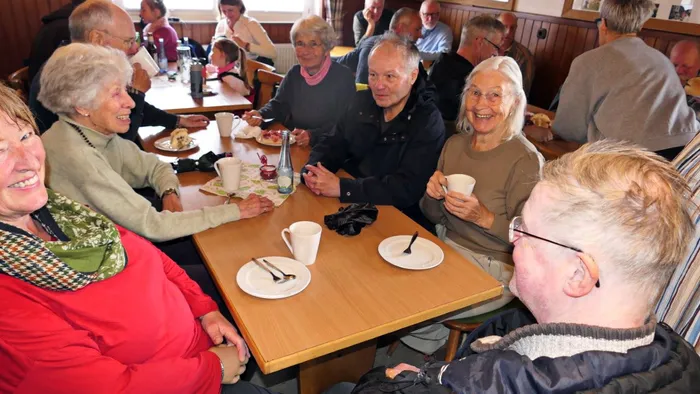 Mehrere ältere Menschen sitzen an einem Tisch in einem hellen Raum zusammen, lächeln und unterhalten sich bei Kaffee und Kuchen.