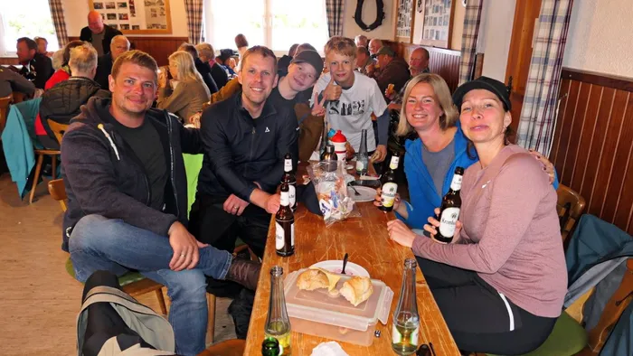 Eine Gruppe von sechs Personen sitzt an einem Holztisch in einem hellen Raum, umgeben von weiteren Menschen. Auf dem Tisch stehen Getränke und eine Brotzeitplatte.