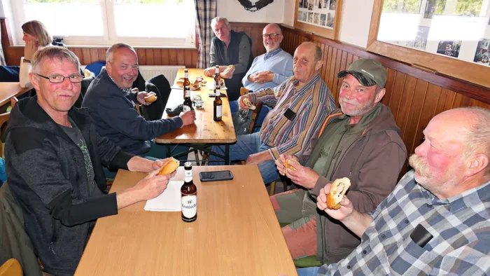 Mehrere Männer sitzen an Holztischen in einem Raum und essen Brötchen, dazu trinken sie Bier. Im Hintergrund sind weitere Personen und eine Fensterfront zu sehen.