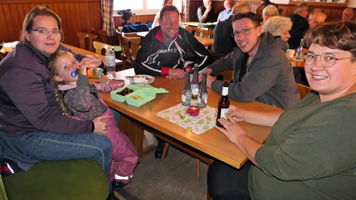 Mehrere Personen sitzen an einem Holztisch in einem hellen Raum und lächeln in die Kamera. Auf dem Tisch stehen Getränke und eine Schachtel mit Gebäck.
