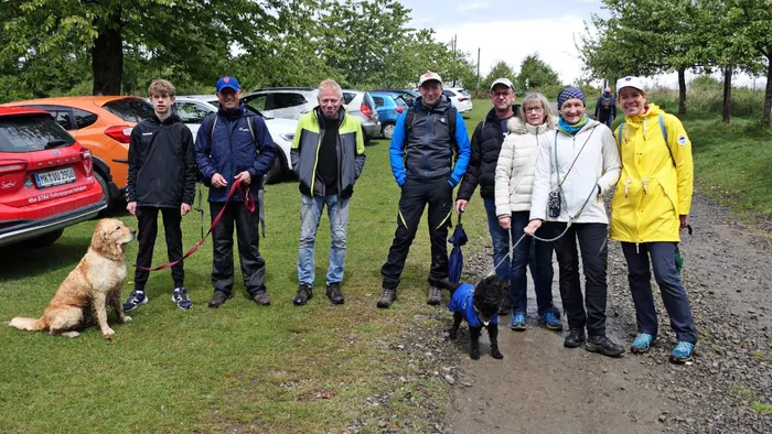 Gruppe von acht Personen mit zwei Hunden steht vor dem Wanderheim Wiehardt an einem bewölkten Tag. Im Hintergrund sind mehrere geparkte Autos zu sehen.