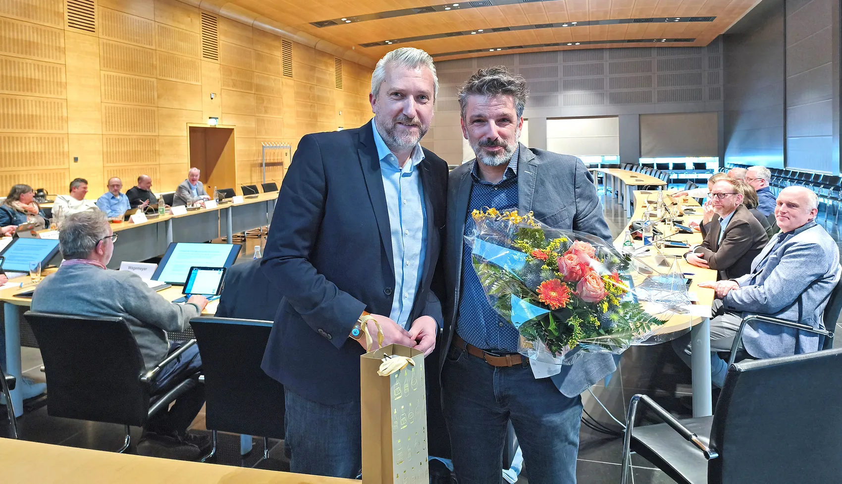 Zwei Männer im Anzug posieren lächelnd im Ratssaal. Der linke Mann, Bürgermeister Sebastian Wagemeyer, überreicht dem rechten Mann, Marcus Müller, eine goldene Geschenkverpackung und einen Blumenstrauß.