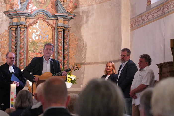Ein Mann spielt Gitarre während einer feierlichen Verabschiedung in einer Kirche vor einem prunkvoll verzierten Altar.