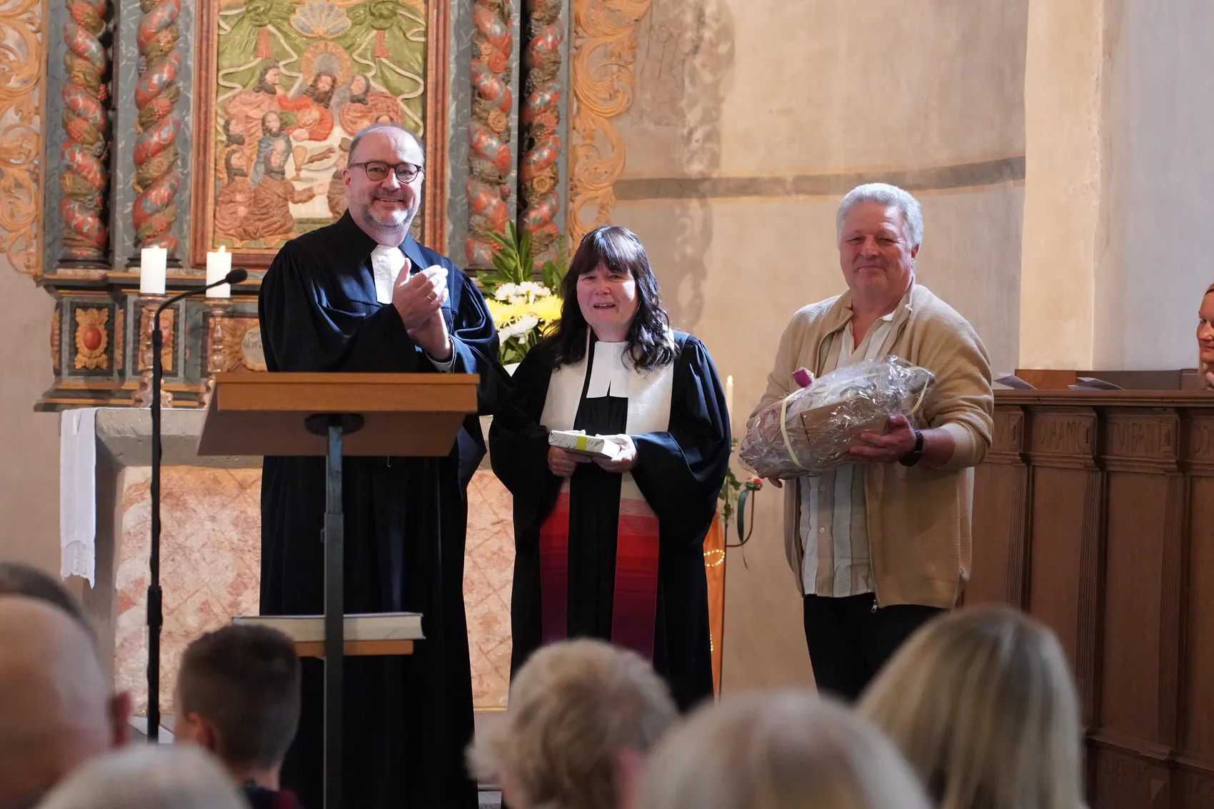 Pfarrerin Anju Laddach steht mit Superintendent Dr. Christof Groote und Wolfgang Laddach, der ein Geschenk hält, vor einem Altar.