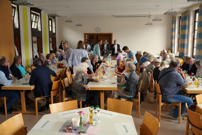 Menschen sitzen an Tischen in einem hellen Raum bei einer feierlichen Verabschiedung im Gemeindehaus.