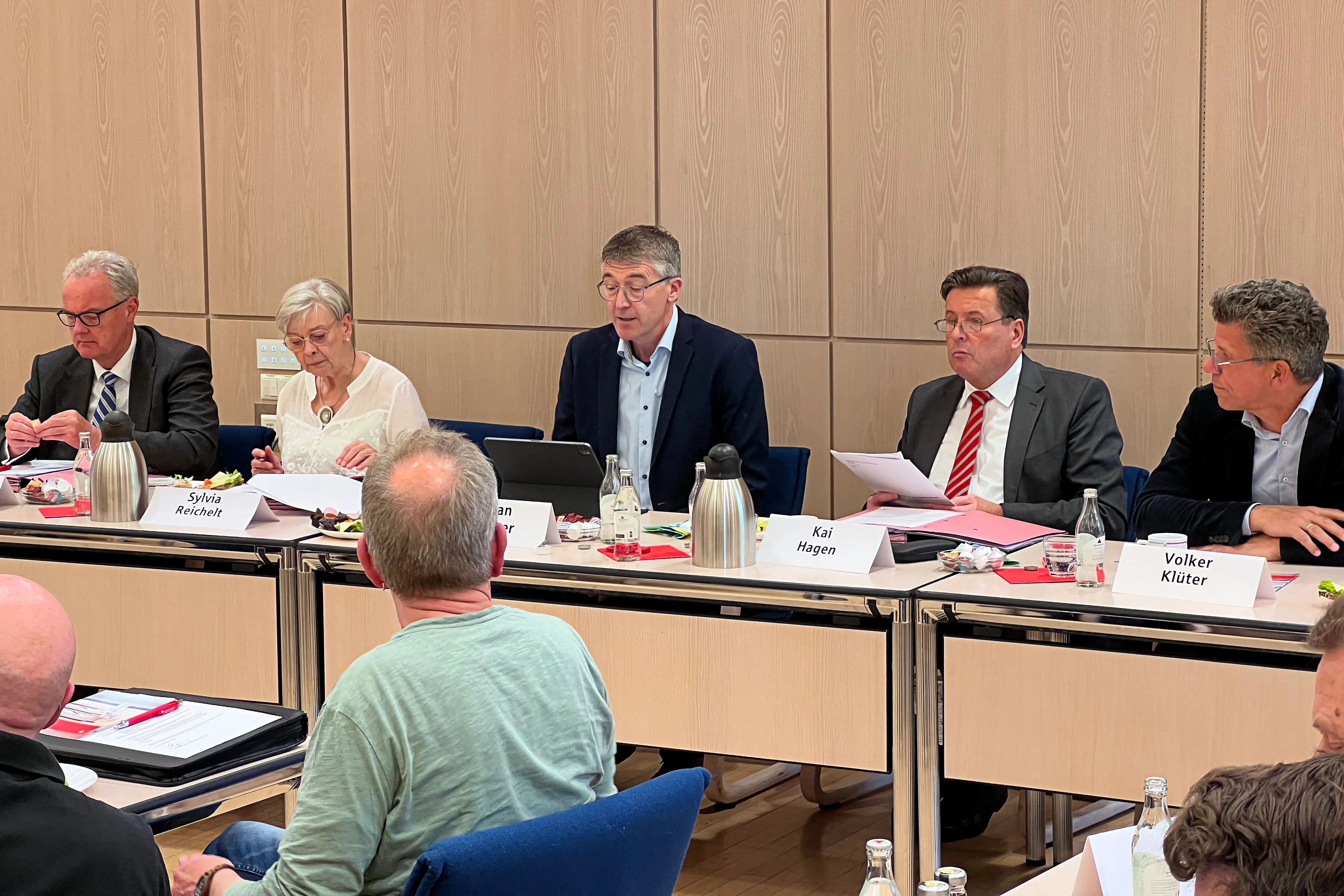 Mehrere Personen sitzen an einem langen Tisch in einem hellen Raum, vermutlich während einer Versammlung der Vereinigten Sparkasse im Märkischen Kreis. Namensschilder vor den Teilnehmern zeigen Sylvia Reichelt, Kai Hagen und Volker Klüter.