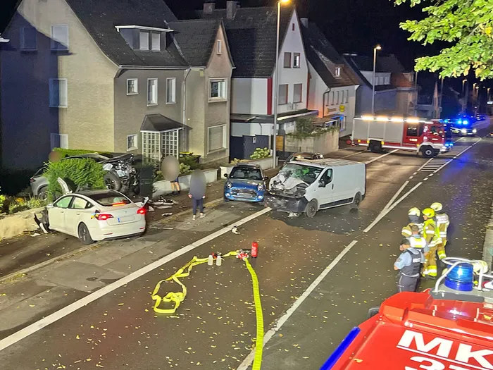 Mehrere beschädigte Fahrzeuge und Einsatzkräfte der Feuerwehr an einem Unfallort vor Wohnhäusern bei Nacht.