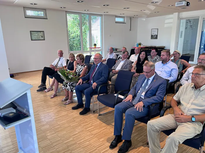 Eine Gruppe von Menschen sitzt in blauen Stühlen in einem hellen Raum bei einer feierlichen Veranstaltung.