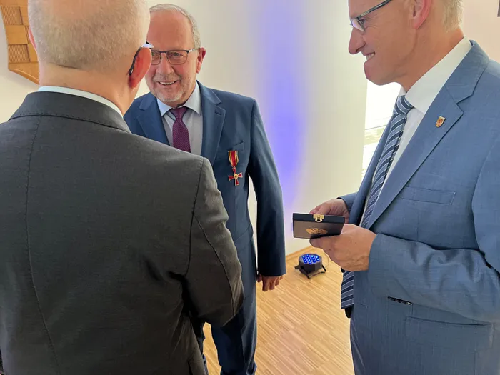 Wolfgang Vöpel lächelt mit dem Verdienstkreuz an seinem blauen Anzug, während ein Mann ihm eine Medaillenschachtel zeigt.