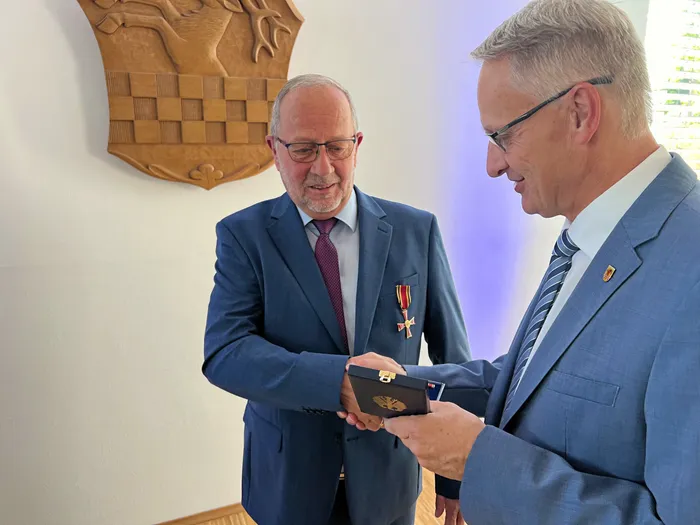 Wolfgang Vöpel nimmt eine Auszeichnung in einer kleinen schwarzen Schachtel entgegen; an seinem Revers ist ein Verdienstkreuz befestigt.