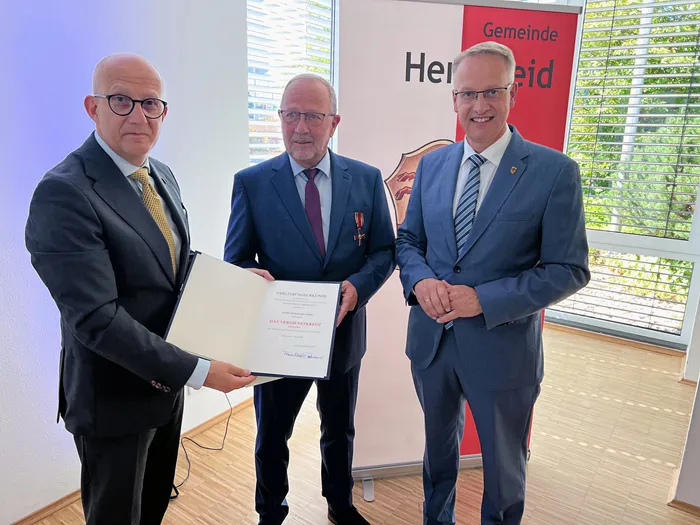 Drei Männer in Anzügen posieren mit einer Urkunde über das Verdienstkreuz vor einem Banner der Gemeinde.