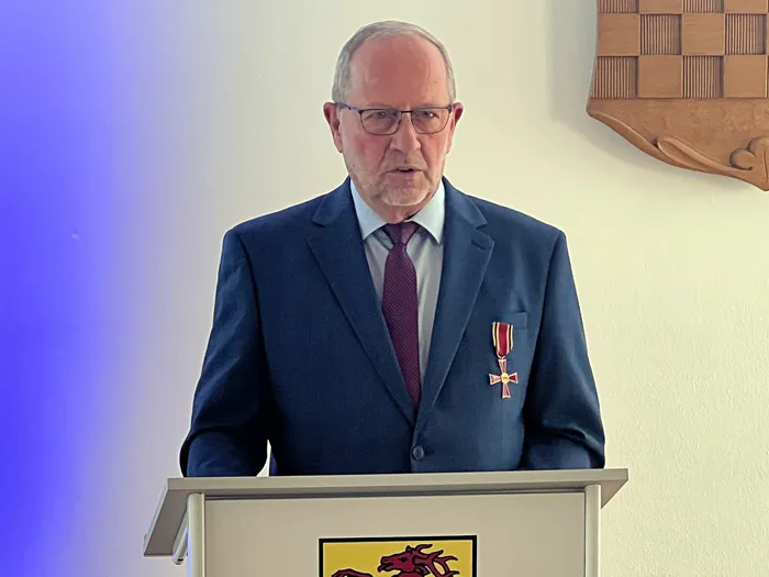 Wolfgang Vöpel mit dem Bundesverdienstkreuz am Revers seines blauen Anzugs hinter einem Rednerpult.