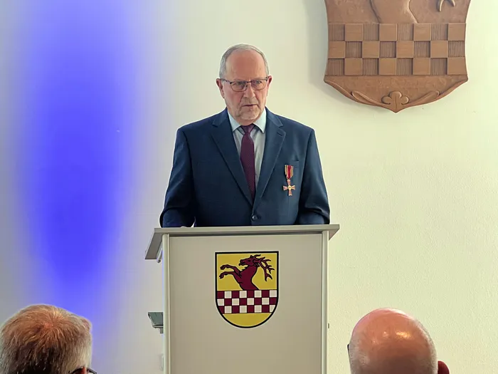 Wolfgang Vöpel steht hinter einem Rednerpult mit einem Wappen und trägt das Bundesverdienstkreuz an seinem Anzug.
