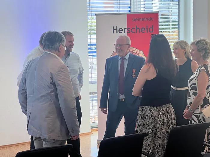 Wolfgang Vöpel steht lächelnd mit dem Verdienstkreuz vor einem Banner der Gemeinde Herscheid inmitten einer Gruppe von Menschen.