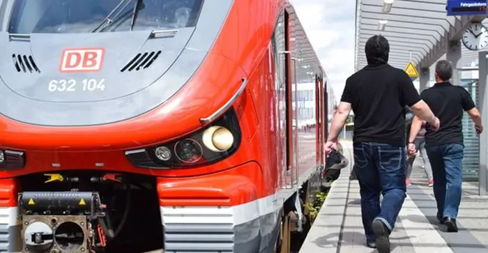 Ein roter DB-Zug steht an einem Bahnsteig, an dem zwei Männer in schwarzer Kleidung entlanggehen.