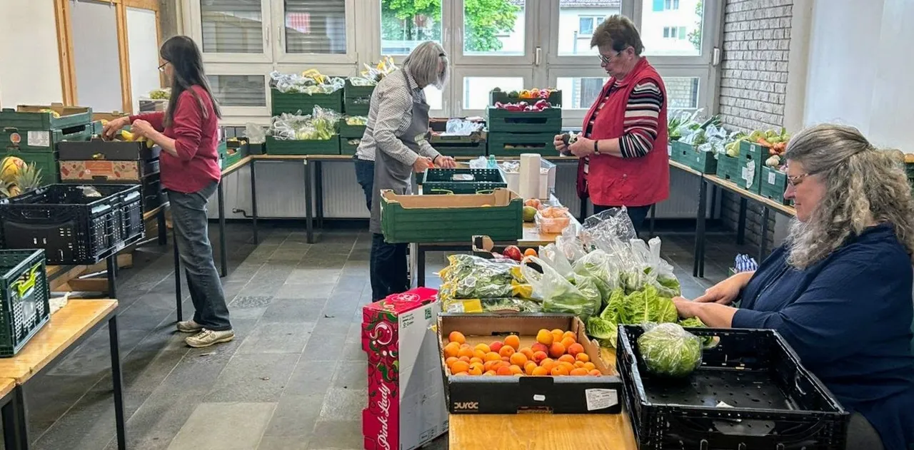 Mehrere Freiwillige sortieren und verpacken frisches Obst und Gemüse in einer großen Vorratskammer. Sie arbeiten an Tischen, die mit Kisten gefüllt sind, und tragen Westen.