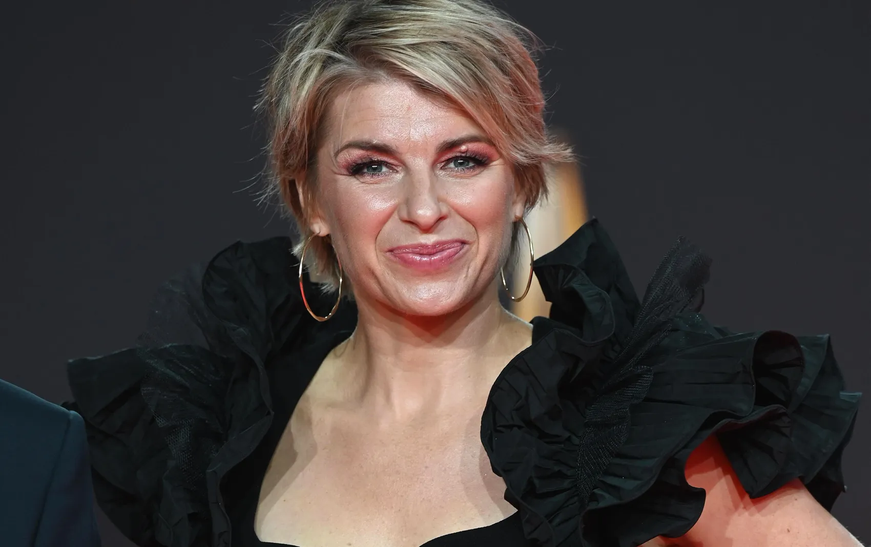 Porträt von Sabine Heinrichs mit blonden Haaren, goldenen Ohrringen und einem schwarzen Kleid mit Rüschen.