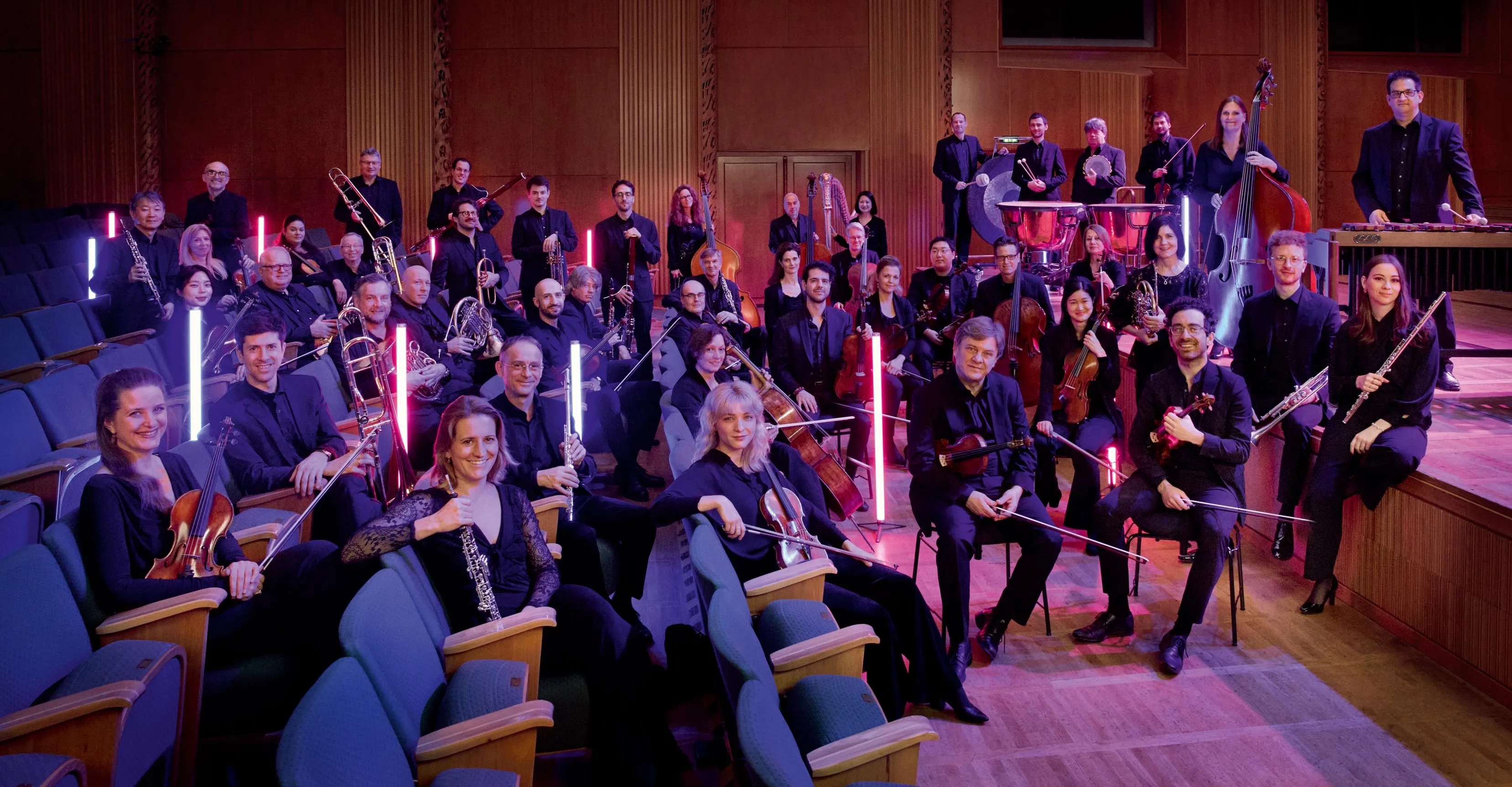 Orchester mit verschiedenen Instrumenten auf einer Bühne, einige Musikerinnen und Musiker lächeln in die Kamera. Im Hintergrund leuchten rote Säulen.
