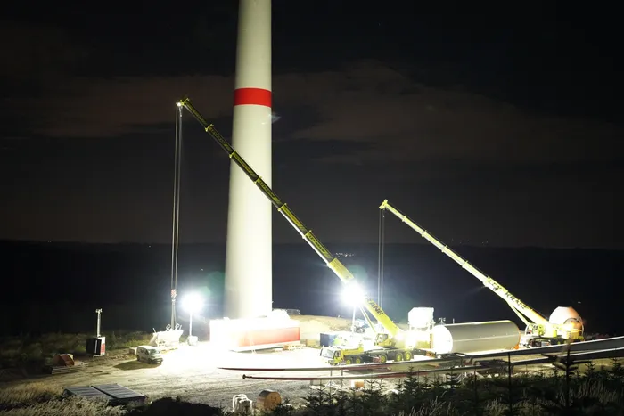 Zwei Mobilkräne bewegen nachts unter Flutlicht Bauteile an der Basis eines weißen Windkraftanlagen-Turms.
