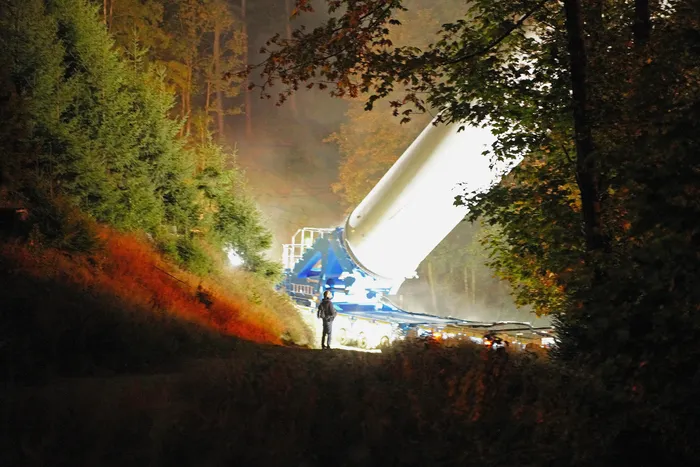 Ein großes weißes Bauteil einer Windkraftanlage wird nachts auf einem blauen Tieflader durch einen Wald transportiert.