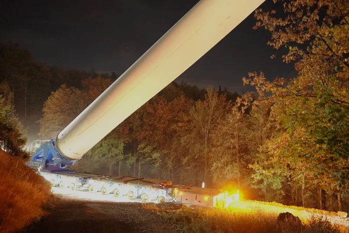 Ein weißer Turmabschnitt einer Windkraftanlage wird auf einem mehrachsigen Tieflader nachts durch einen Wald transportiert.