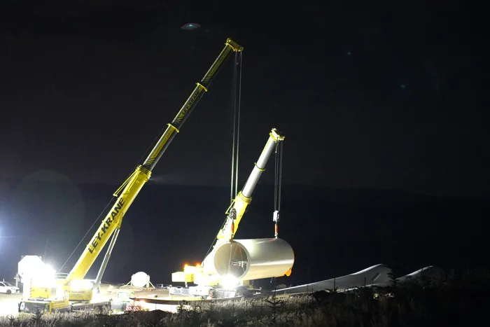 Zwei Kräne heben bei Nacht ein großes, zylindrisches Turmelement für eine Windenergieanlage.