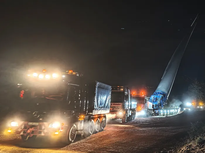 Ein Lastwagen-Tross transportiert nachts ein langes Rotorblatt einer Windkraftanlage auf einer unbefestigten Straße.