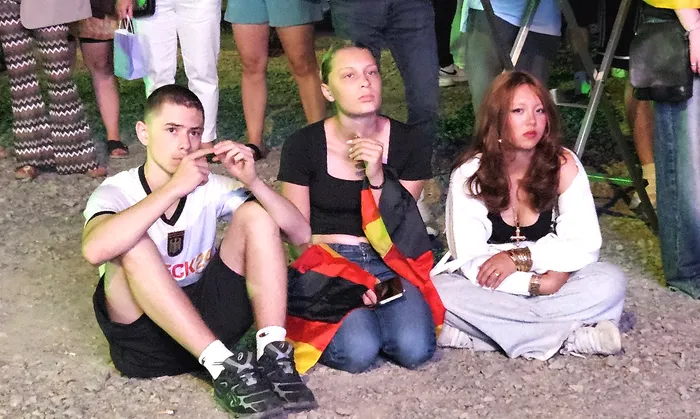 Drei junge Menschen sitzen auf dem Boden, die Gesichter zeigen Enttäuschung und Sorge. Ein Fan hält eine Deutschlandflagge, während die anderen mit den Händen ihre Gesichter bedecken.
