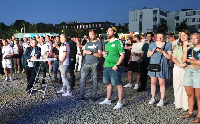 Eine große Menschenmenge schaut auf Bildschirme, einige halten Getränke in der Hand. Im Hintergrund sind moderne Gebäude und Bäume zu sehen.