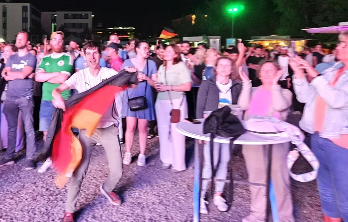 Ein Mann schwenkt eine Deutschlandflagge inmitten einer jubelnden Menschenmenge bei einer Public-Viewing-Veranstaltung. Im Hintergrund sind weitere Fans und eine zweite, kleinere Deutschlandflagge zu sehen.