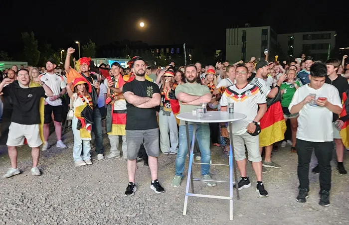 Eine große Menschenmenge jubelt und schaut auf Bildschirme, viele tragen Deutschland-Trikots und schwenken Flaggen. Einige stehen an Tischen, andere halten ihre Handys hoch.
