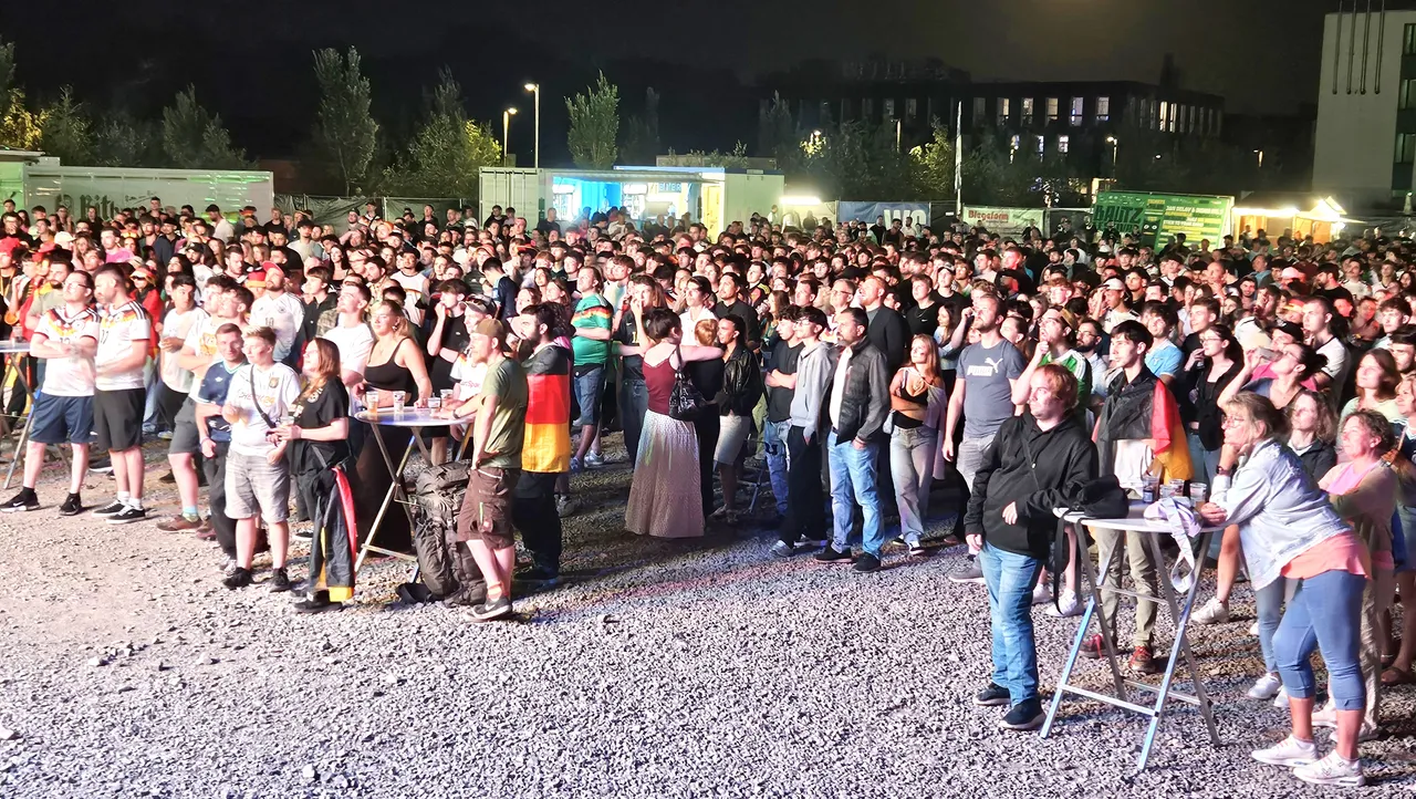 Große Menschenmenge beim Public Viewing eines Fußballspiels, viele halten Deutschlandflaggen hoch. Im Hintergrund sind Buden und ein beleuchtetes Gebäude zu sehen.