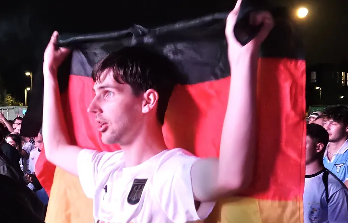 Ein Mann hält eine deutsche Flagge hoch, während er in die Ferne schaut. Im Hintergrund sind weitere Menschen zu sehen.