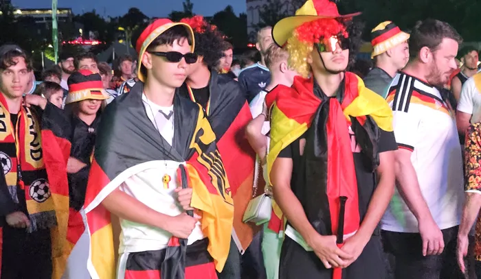 Deutsche Fußballfans mit Deutschlandflaggen und in Nationalfarben gekleidet stehen während eines Public Viewings mit ernstem Gesichtsausdruck da. Einige tragen Sonnenbrillen und einen Hut mit rotem Haar. Im Hintergrund sind weitere Fans zu sehen.