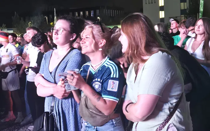 Eine Gruppe von Menschen schaut aufgeregt nach oben, einige mit verschränkten Armen. Im Vordergrund trägt eine Frau ein Trikot der deutschen Nationalmannschaft. Im Hintergrund sind weitere Zuschauer und Gebäude zu sehen.
