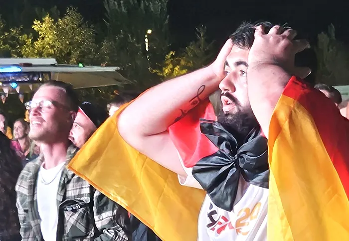 Ein Mann mit einer Deutschlandflagge um die Schultern hält sich die Hände an den Kopf, während im Hintergrund ein anderer Mann mit geschlossenen Augen zu sehen ist. Die Stimmung bei den Fans nach dem enttäuschenden Spiel der deutschen Nationalmannschaft ist spürbar.